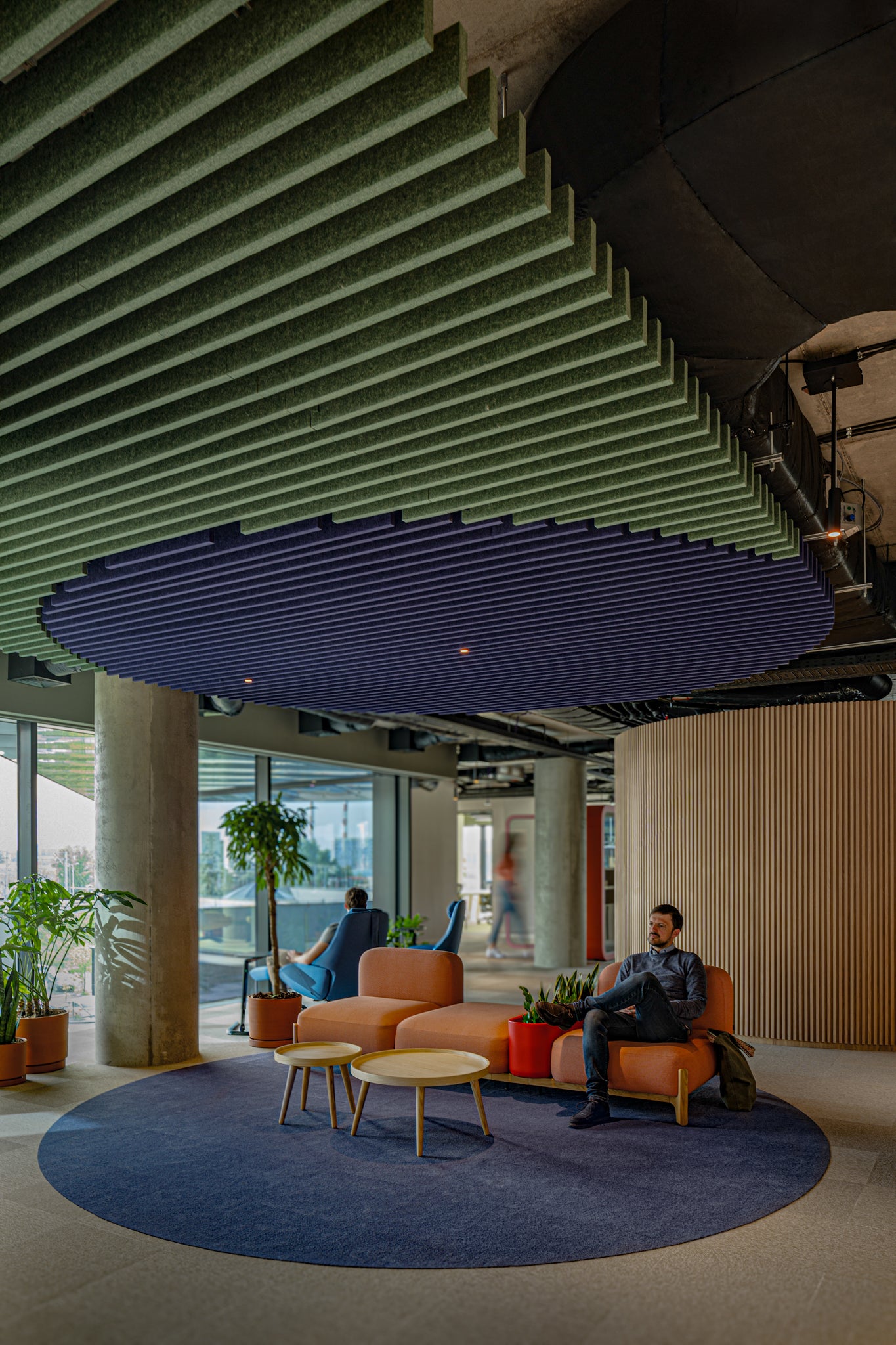 Deckenbaffel Sraight Modular – Deckenbaffel – Loungebereich mit orangefarbenem Sofa, zwei runden Holztischen und blauen Teppich vor Fensterfront. Pflanzen und Personen im Hintergrund.