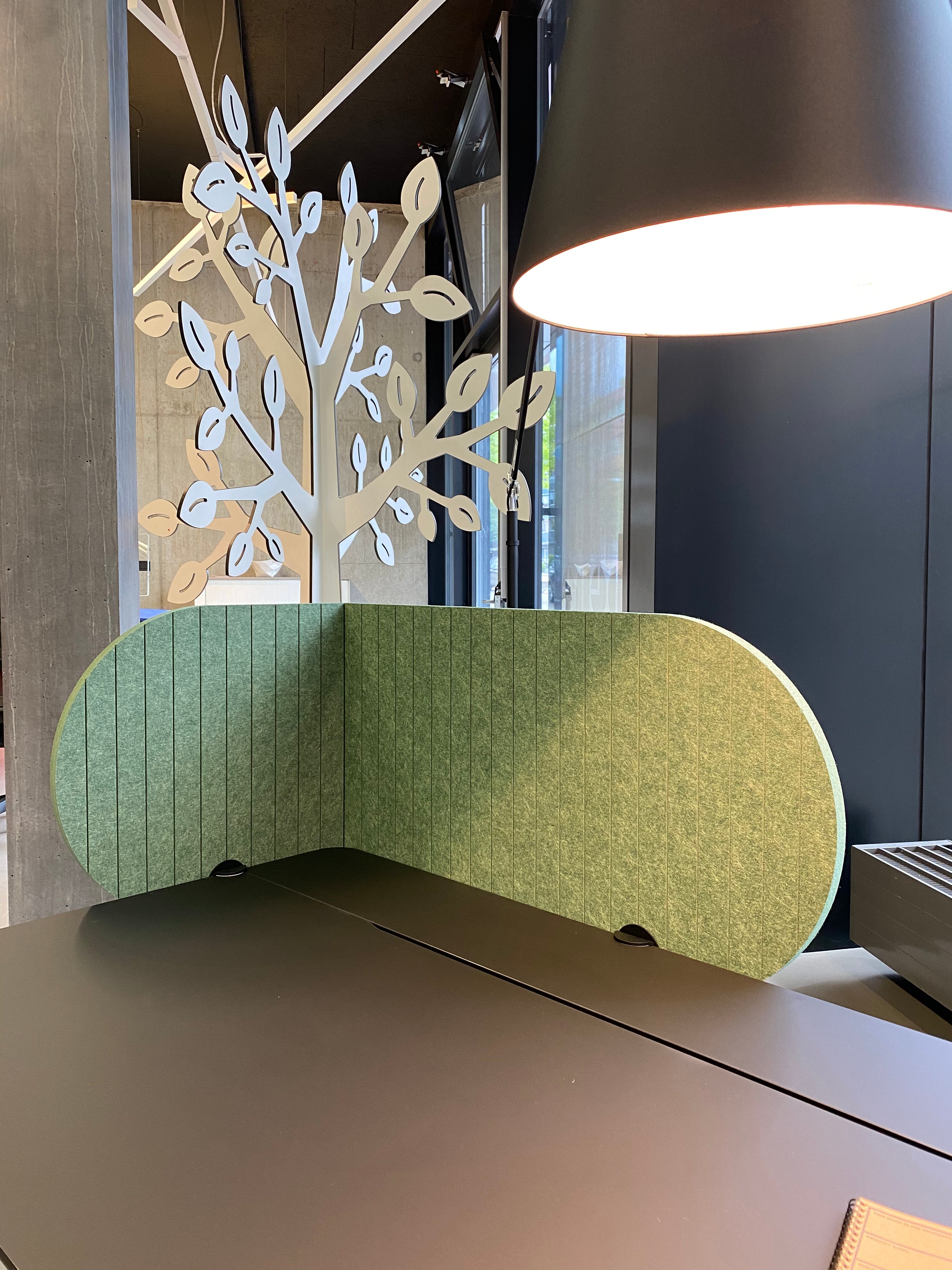 Arbeitsplatztrenner Half Moon – Akustische Tischtrenner – Grüne Trennwand aus Akustikmaterial auf schwarzem Tisch, mit beleuchtetem Lampenschirm im Hintergrund und dekorativem Baummotiv.