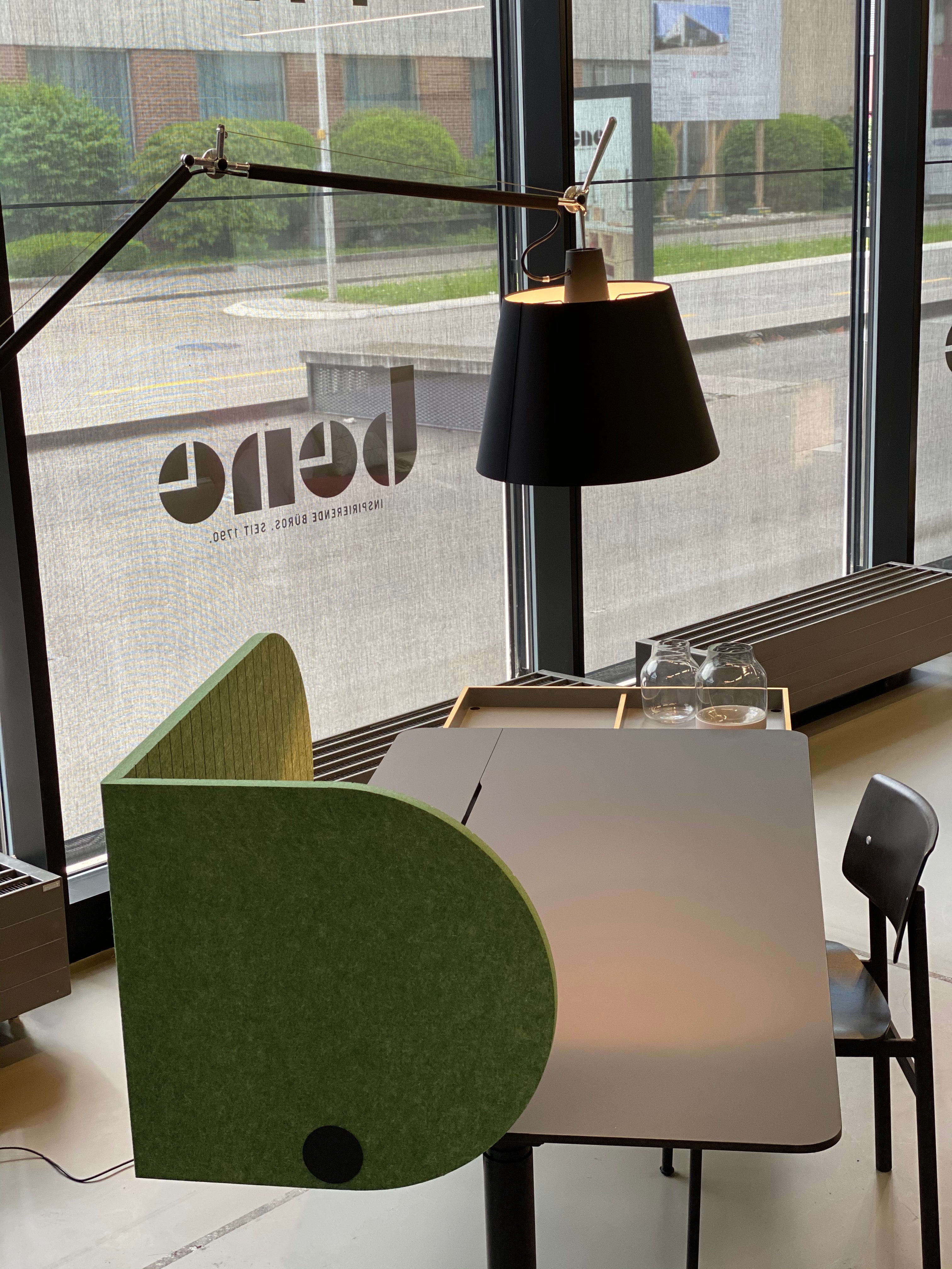 Arbeitsplatztrenner Half Moon – Akustische Tischtrenner – Grüner Trennwand-Element neben einem schlichten Tisch, schwarzweißer Lampe und Stuhl im modernen Büroambiente.