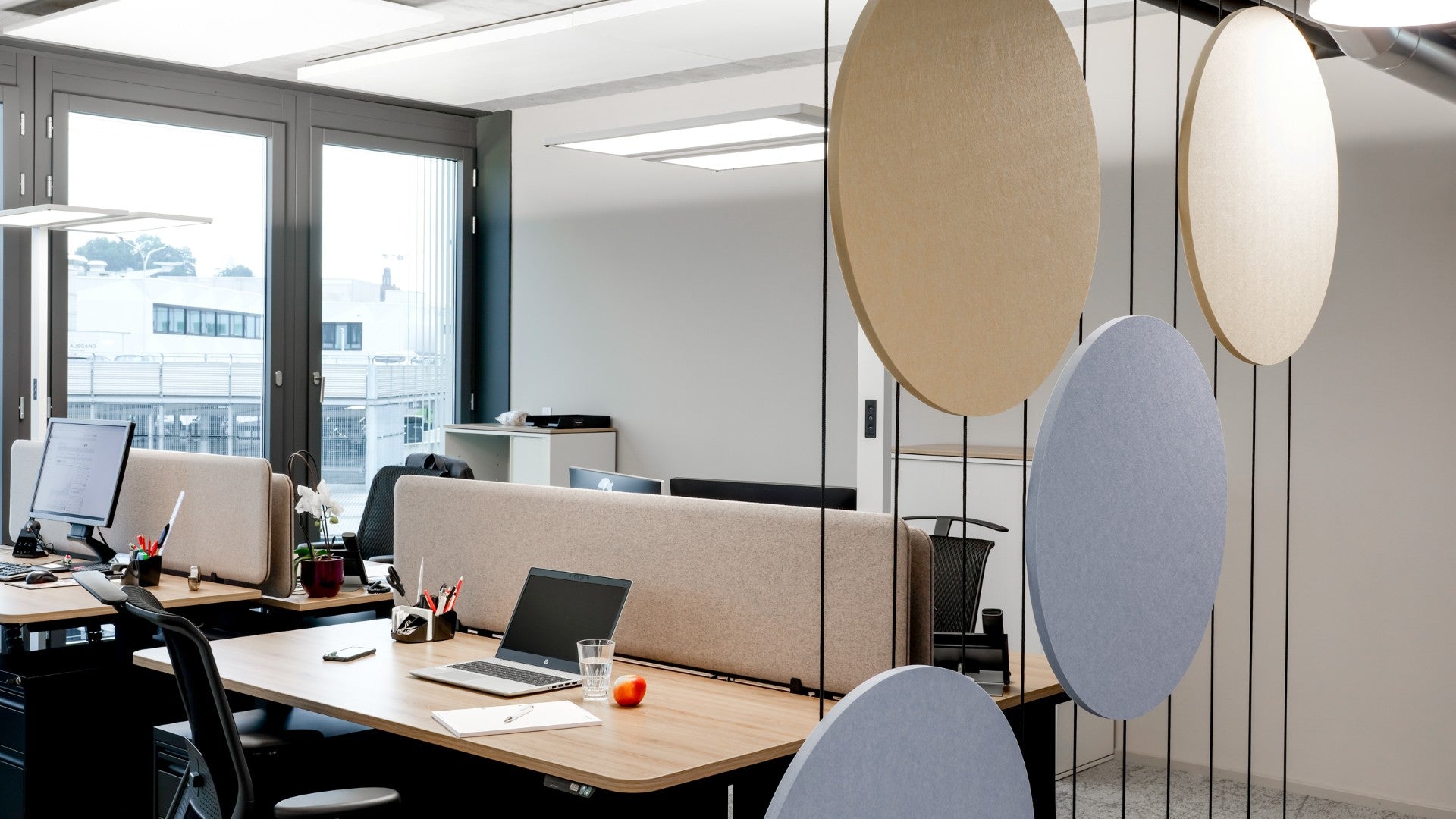 Gespannte Trennwand Helios – Akustische Raumtrenner – Moderne Büroumgebung mit Tisch, Monitor, Akustikpaneelen und Pflanzen. Helle Farben und viel Tageslicht.