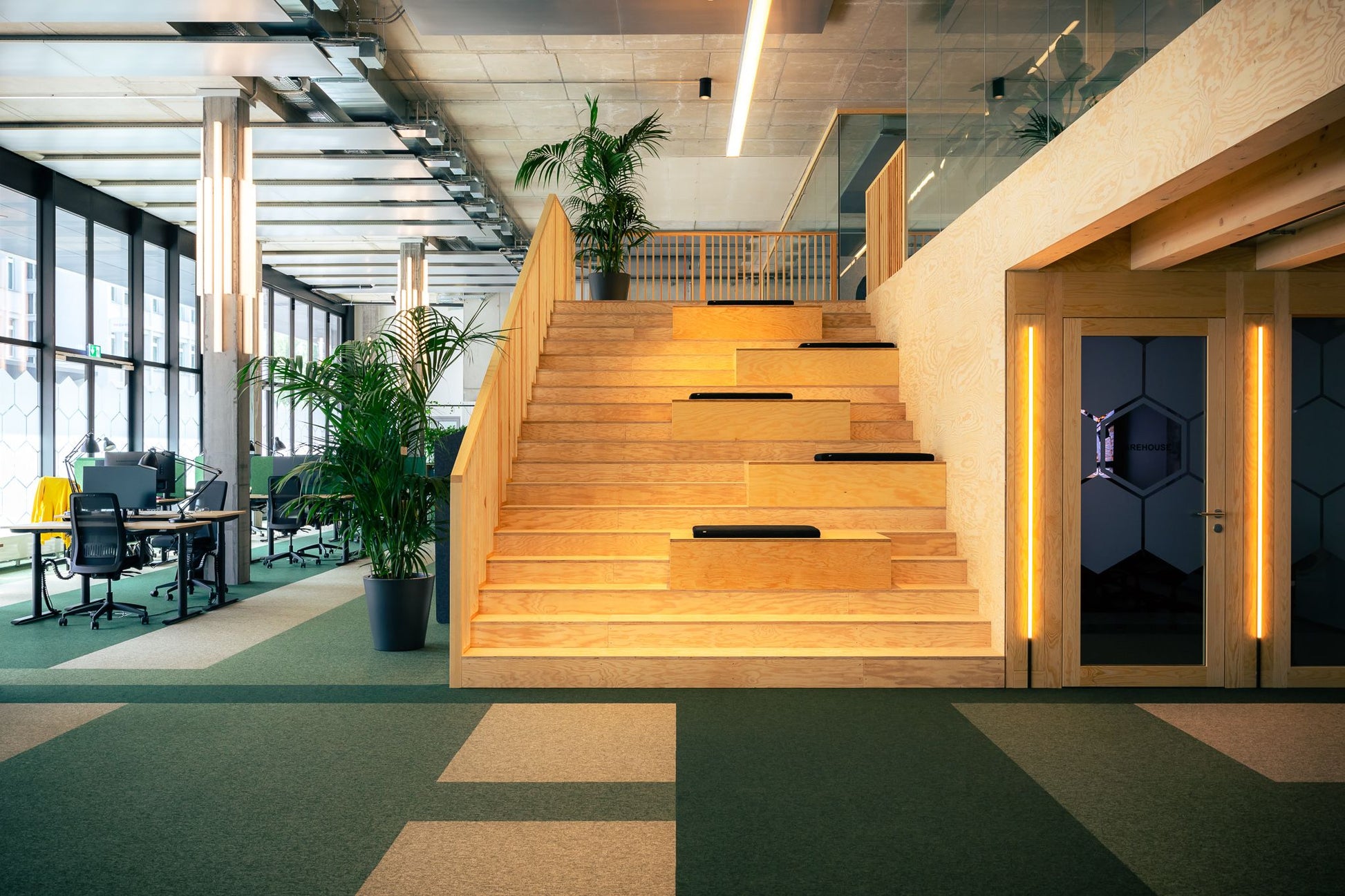 Arbeitsplatztrenner Supra – Akustische Tischtrenner – Holztreppe mit Beleuchtung im modernen Büro, sichtbare Schreibtische und Pflanzen im Hintergrund.