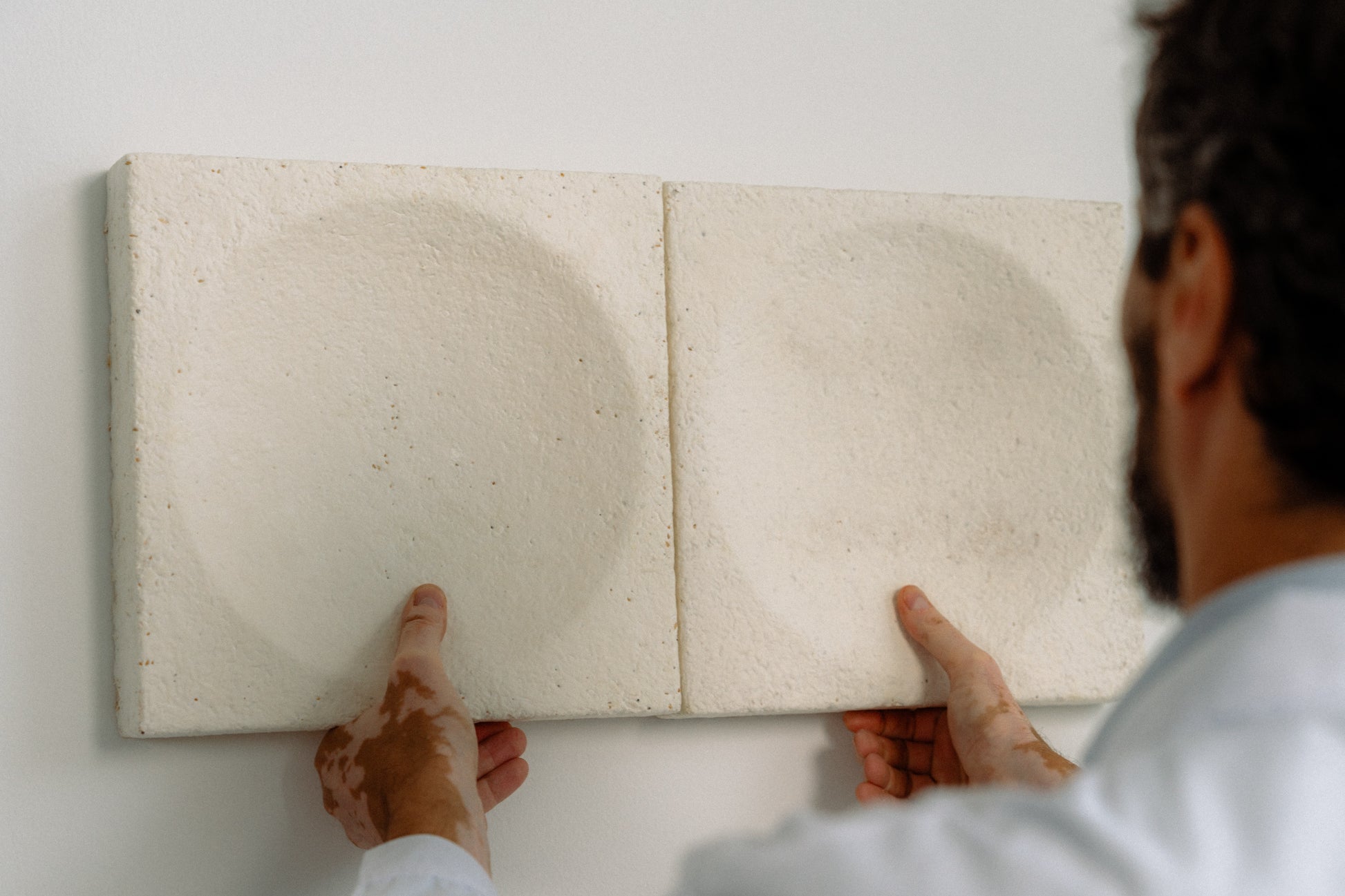Archisonic Mycelium Wandfliesen – Akustische Wandverkleidung – Zwei quadratische Wandpaneele in weißer Farbe werden von einer Person an die Wand gehalten.