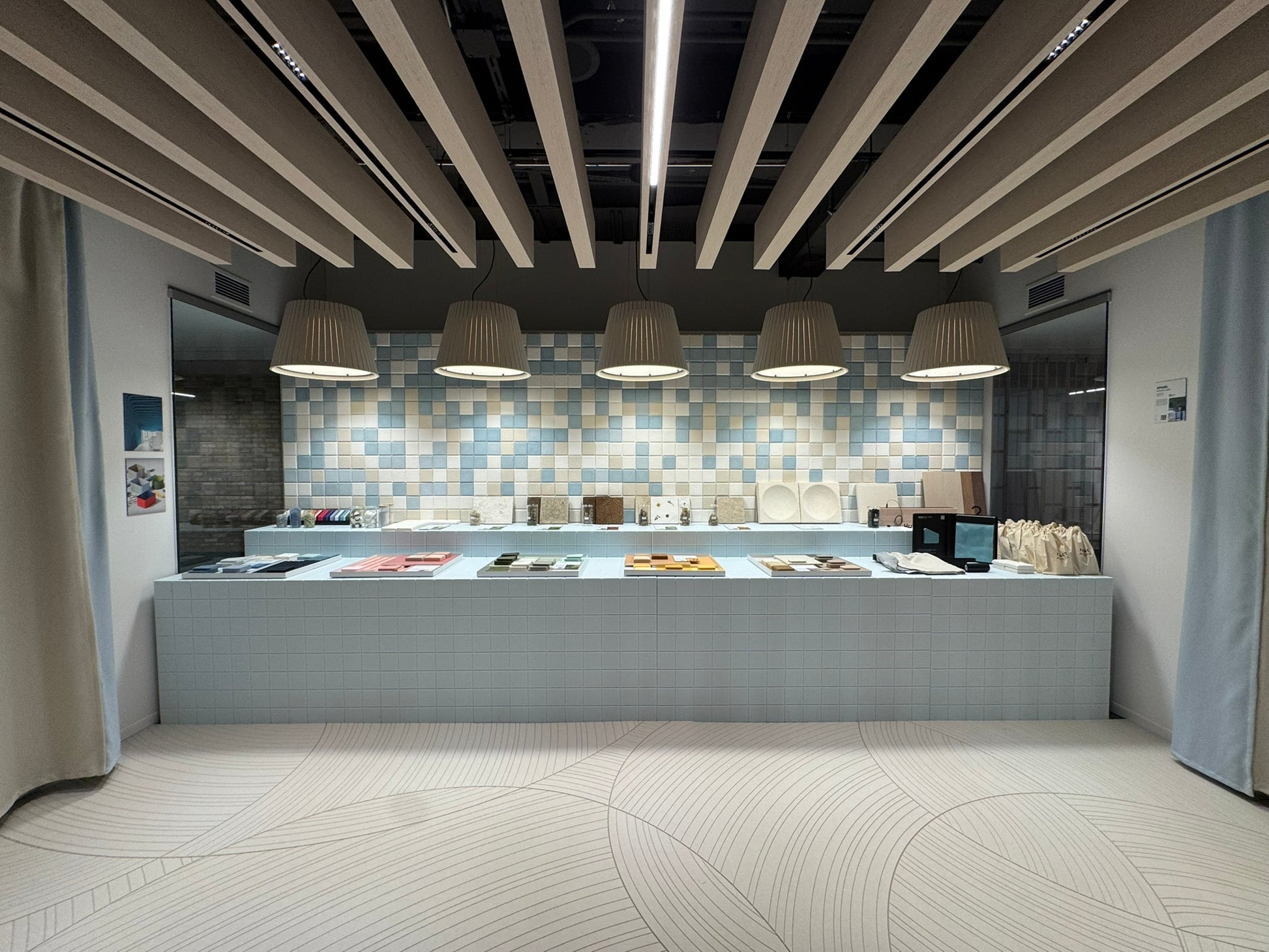 Archisonic Cotton Mosaik – Akustische Wandverkleidung – Moderne Ausstellung mit hellblauem Kissentisch, dekorativen Wandfliesen in Blau und Beige sowie hängenden Lampen.