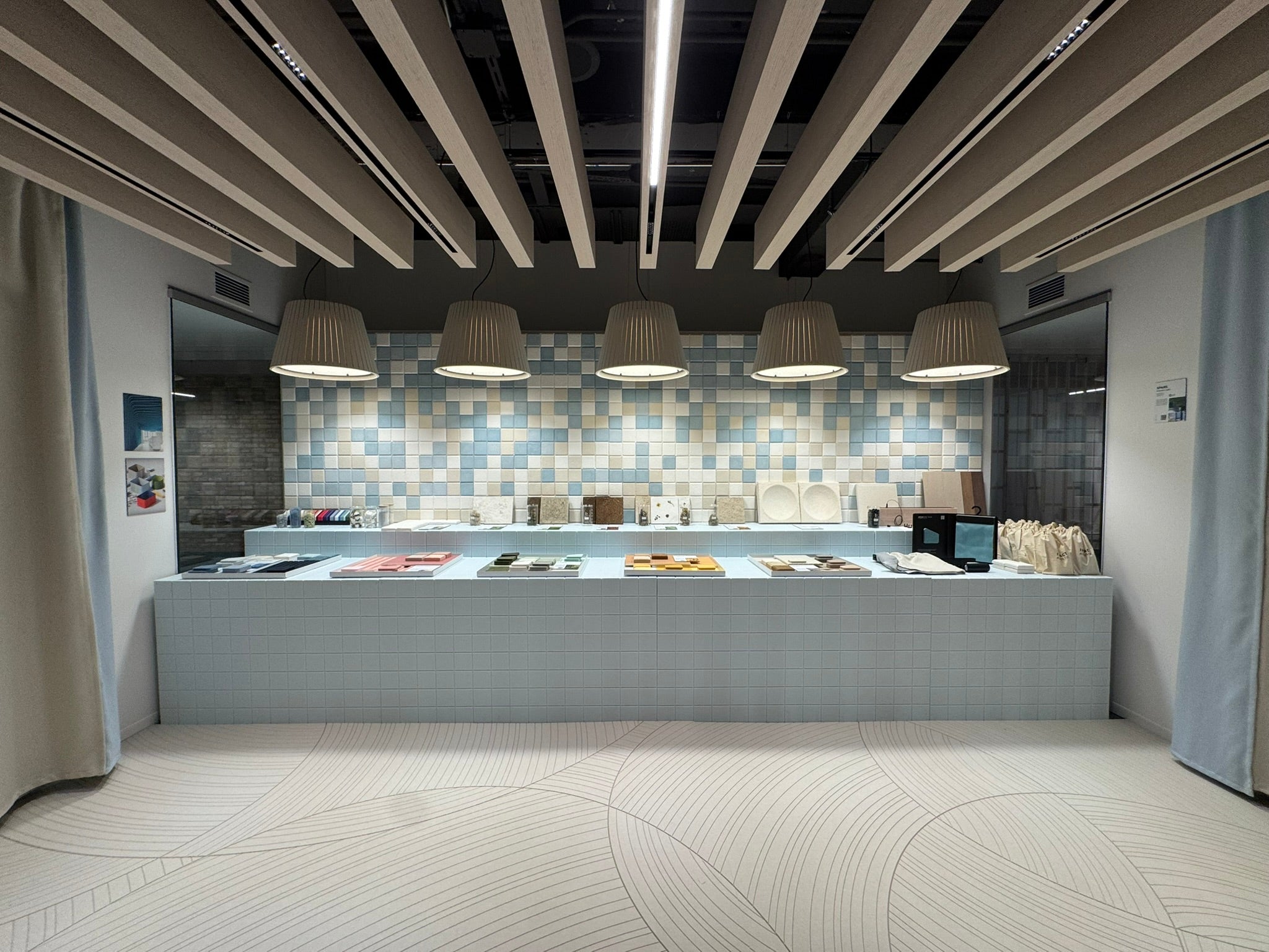Archisonic Cotton Mosaik – Akustische Wandverkleidung – Moderne Ausstellung mit hellblauem Kissentisch, dekorativen Wandfliesen in Blau und Beige sowie hängenden Lampen.