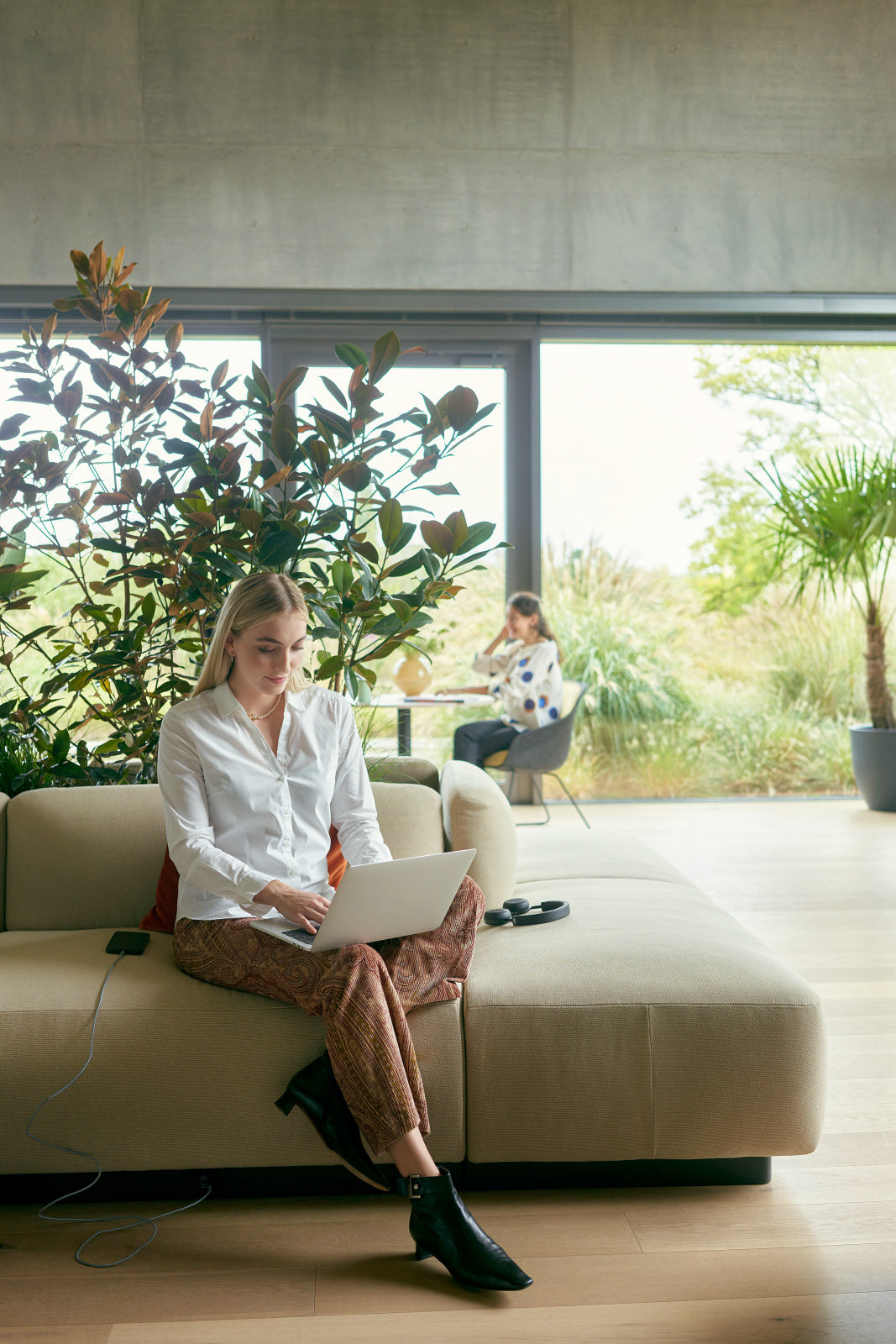 se:living | plant bench – Sofa – Person sitzt auf einem beigen Sofa und arbeitet am Laptop, umgeben von Pflanzen und grossen Fenstern.