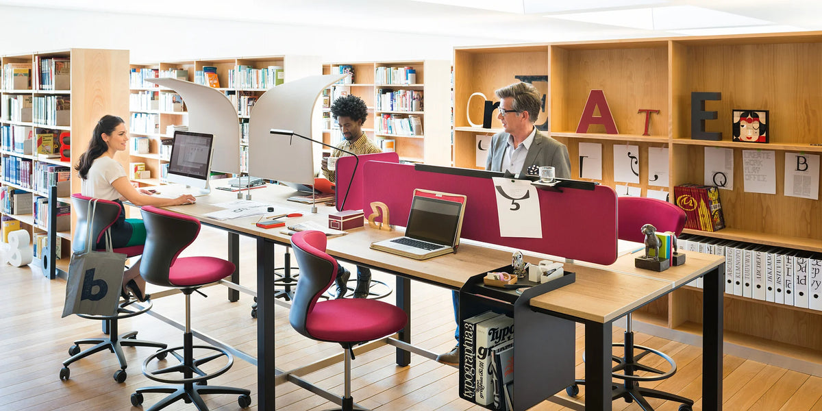easy screen silent – Schreibtischaufsatzelement – Drei Personen arbeiten an einem gemeinsamen Tisch mit pinken Bürostühlen in einem modernen Büro mit Bücherregalen.