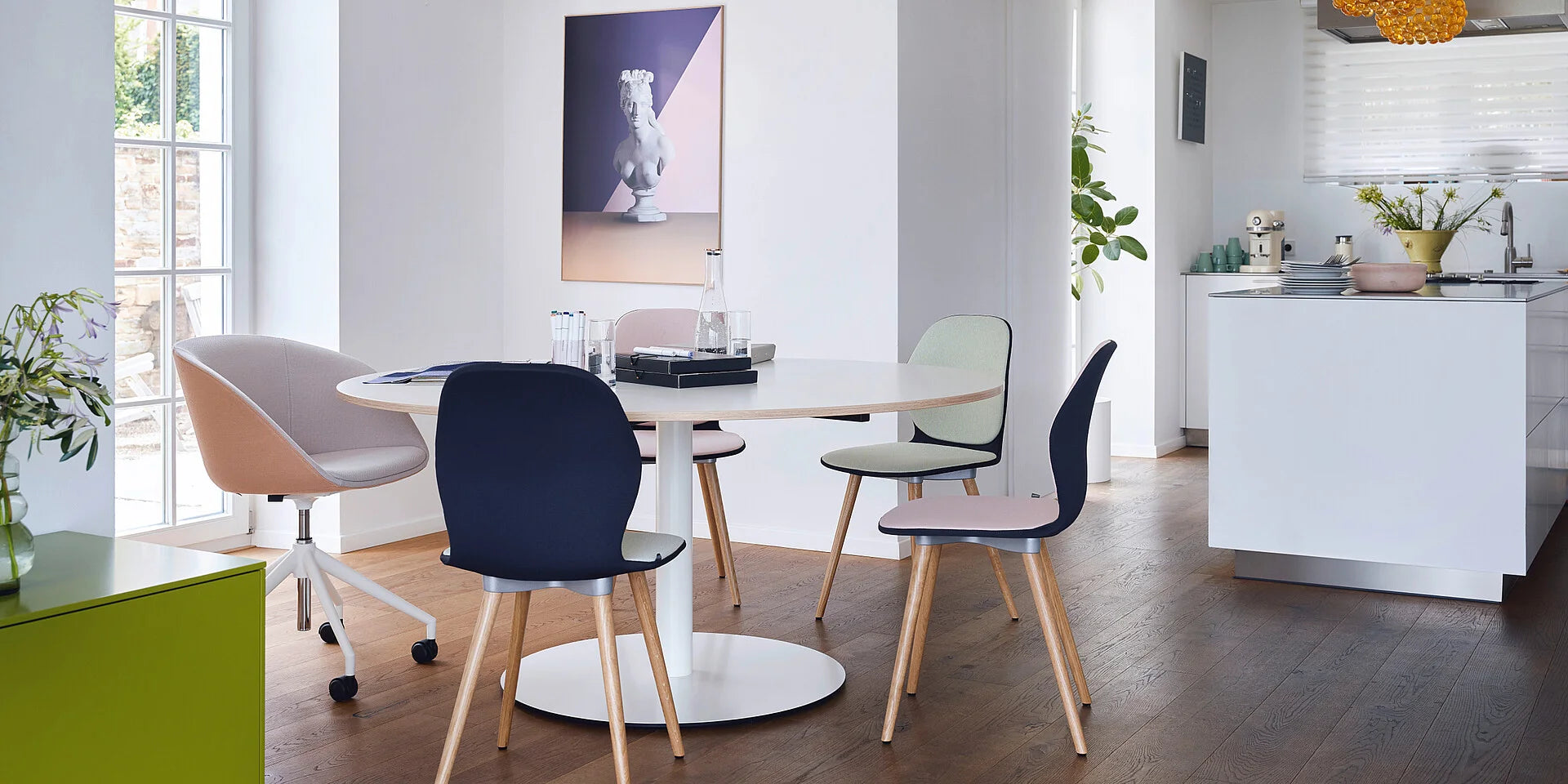se:assist – Loungetisch – Runder Tisch mit vier Stühlen in verschiedenen Farben. Helle Umgebung mit Pflanzen und Küchenbereich im Hintergrund.