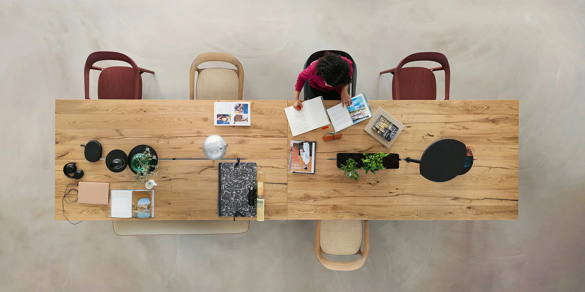 se:café table – Team-Tisch – Holztisch mit mehreren Stühlen, Bücher und Pflanzen auf der Tischfläche. Person blättert in einem Buch.
