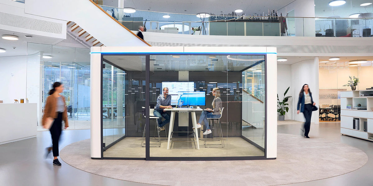 Meetingbox se:cube max: Glasbasiertes Besprechungszimmer mit einem Tisch, zwei Stühlen und Bildschirm, umgeben von offener Bürofläche.