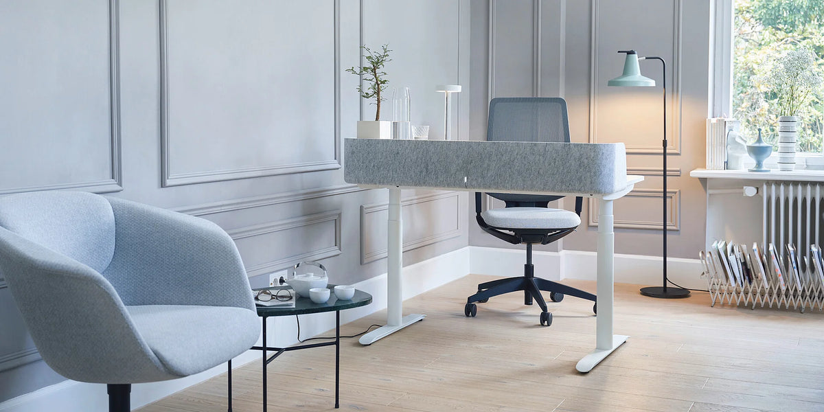 Elektrisch höhenverstellbarer Schreibtisch se:desk home: Grauer Schreibtisch mit Stofftrennwand, schwarzer Bürostuhl, grauer Sessel und Stehlampe in modernem Raum.