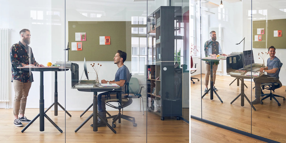 se:lab e-desk A – elektrisch höhenverstellbarer Schreibtisch – Zwei Personen in modernem Büro mit Stehtisch und Schreibtisch. Links ein Stehtisch, rechts ein Schreibtisch mit Computer.