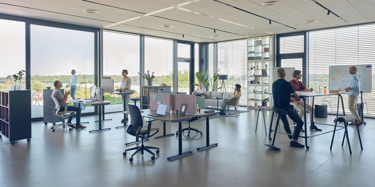 se:lab e-desk – elektrisch höhenverstellbarer Schreibtisch – Moderne Büroumgebung mit verschiedenen Arbeitsplätzen. Personen interagieren an Stehtischen und auf Bürostühlen.