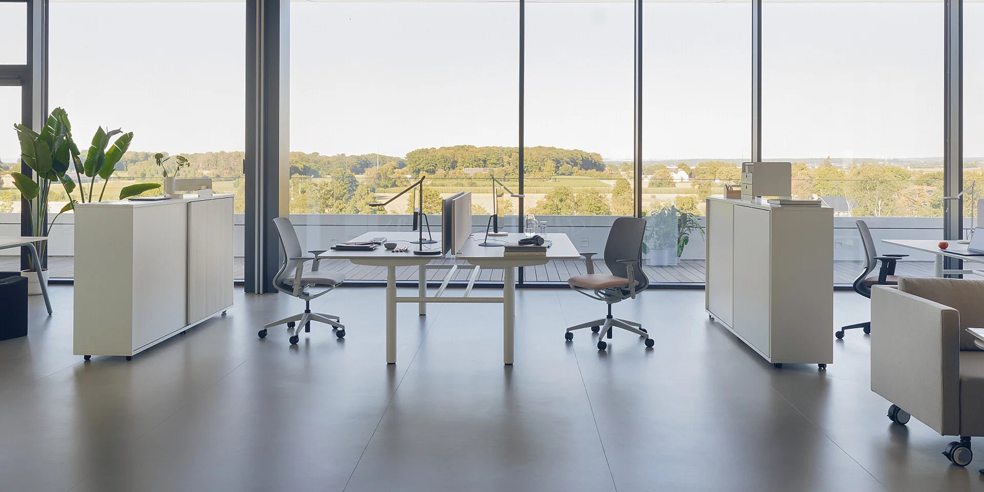 se:lab twin – elektrisch höhenverstellbarer Doppelschreibtisch – Modernes Büro mit zwei Schreibtischen, ergonomischen Stühlen und Pflanzen im Hintergrund. Grosse Fenster mit Aussicht.