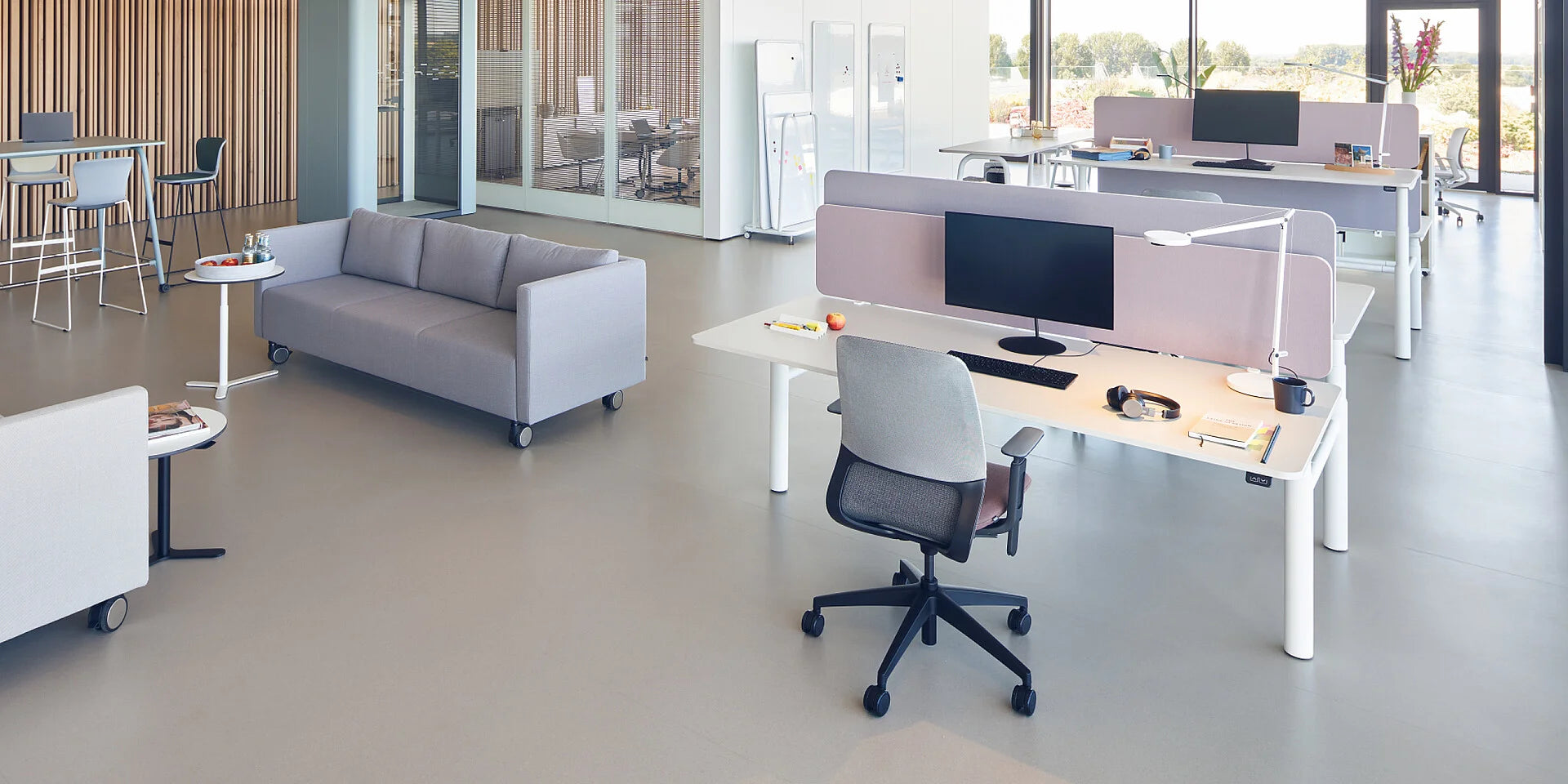 se:lab twin – elektrisch höhenverstellbarer Doppelschreibtisch – Moderne Büroumgebung mit hellgrauem Sofa, weissen Tischen und Stühlen, Arbeitsplätzen mit Monitoren und Trennwänden.