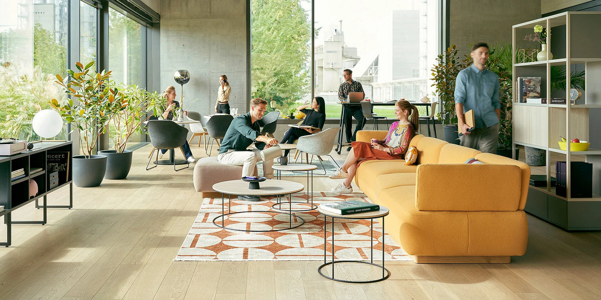 se:living – Loungetisch – Moderne Bürolandschaft mit orangefarbener Couch, runden Tischen, Pflanzen und Personen in verschiedenen Arbeitspositionen.