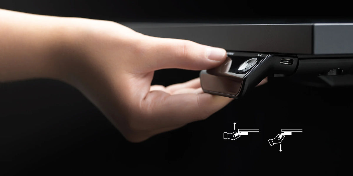 temptation speed – elektrisch höhenverstellbarer Schreibtisch – Hand betätigt einen Mechanismus an einem Schreibtisch zur Einstellung der Höhe. Im Hintergrund ist ein dunkler Farbton.