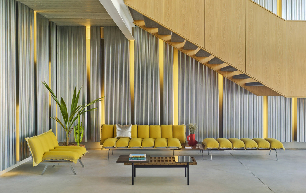 Sancal Next Stop Bank in Gelb, über ein Tischteil mit einer Sitzkomposition verbunden, in einer Halle mit Holztreppe