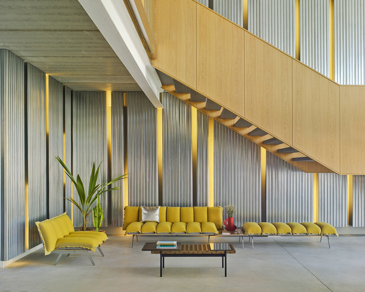 Sancal Next Stop Sessel in Gelb neben einem langen Next Stop Sofa mit Bank, in einer hohen Halle mit Wellblechwand und Holztreppe